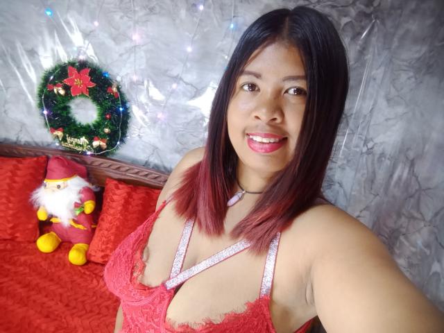 MariahSquirting69, de olhos castanhos, sorrindo, usando um sutiã de renda vermelho, mostrando seus seios, com batom rosa e cabelo castanho escuro, em uma cama vermelha, com uma guirlanda de Natal e uma boneca do Papai Noel..