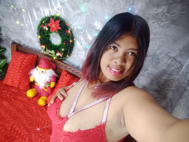 MariahSquirting69 with medium tits in red lace lingerie, smiling, holding up phone, black hair with red tips, Christmas decor, Santa doll, wreath, and lights in the background.