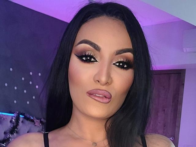 NovaDiamond has dark, wavy hair, heavy black eyeliner, and pink, glossy lips. She's wearing a black top and has a neutral expression with purple lighting in the background.