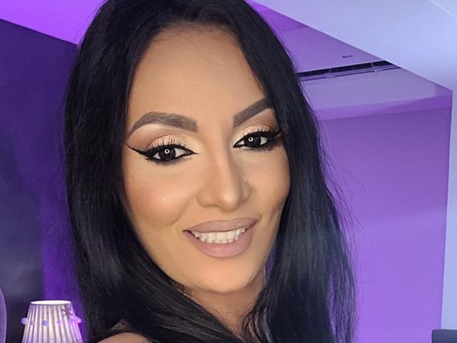 NovaDiamond's face with long black hair, thick eyebrows, winged eyeliner, and a smile showing her teeth. She's wearing a black top, and the purple lighting is behind her.