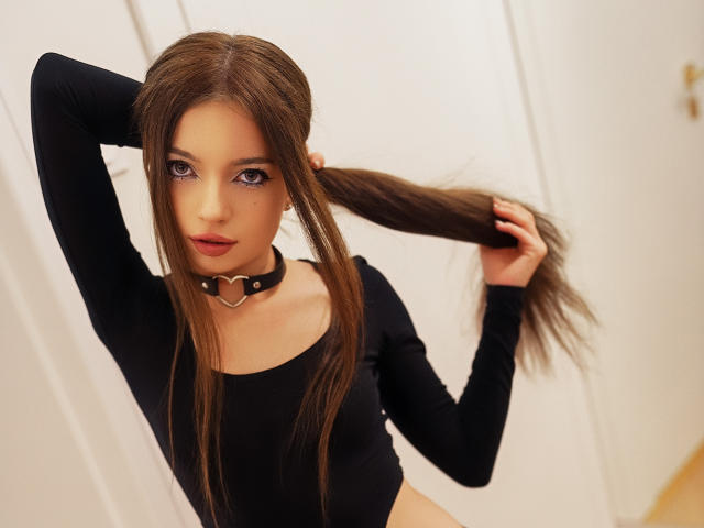 GraceGold with long brown hair, wearing a black choker with a heart-shaped metal ring, and a black long-sleeve top, holding her hair with one hand.