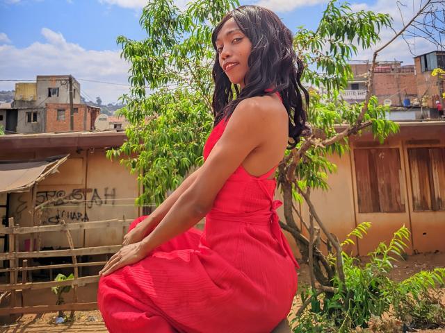 VictorineStone with dark brown skin and long black hair, wearing a red dress, sitting on a rooftop, looking back, hands on her knee, green tree behind her.