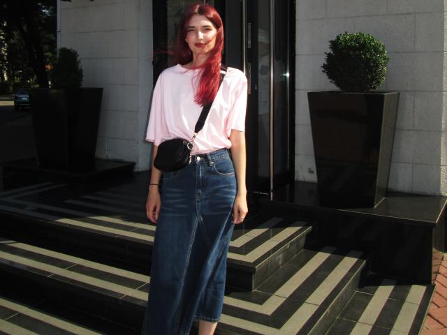 LisaDarcy with red hair, pink shirt, blue high-waisted skirt, black crossbody bag, standing on striped black-and-white steps, smiling, wearing red lipstick, hands by her sides.