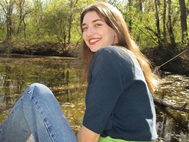LisaDarcy with long brown hair, smiling, wearing a dark blue shirt and blue jeans, sitting by a creek, showing her white ass slightly, in a sunny forest.
