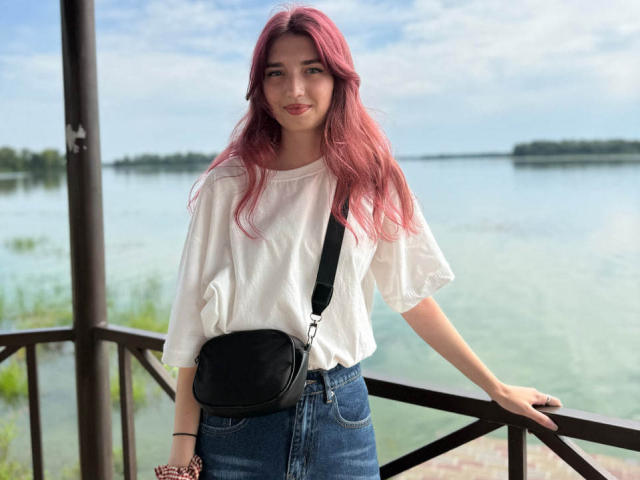 LisaDarcy with pink hair, white loose shirt, blue jeans, black crossbody bag, standing on wooden balcony, smiling, holding wrist with red bracelet, blue sky, cloudy, water behind.