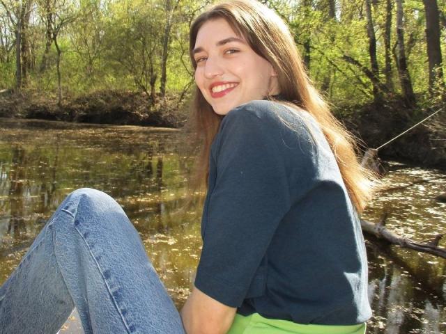 LisaDarcy with long brown hair, smiling, wearing a dark blue shirt and green pants, sitting by a shallow, sunlit creek with her knees up and legs crossed.