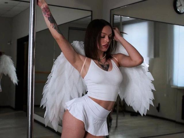 PrettyEllie-hot stands in a room with a mirror and pole, wearing white angel wings, crop top, and skirt. She has light skin, brown hair, and a tattoo on her right arm. Her right arm is raised, gripping the pole, while her left hand touches her hair. She has a heart necklace and a confident, slightly sensual expression. The white wings are textured, and her top and skirt are simple and white. The image has a realistic, modern style.