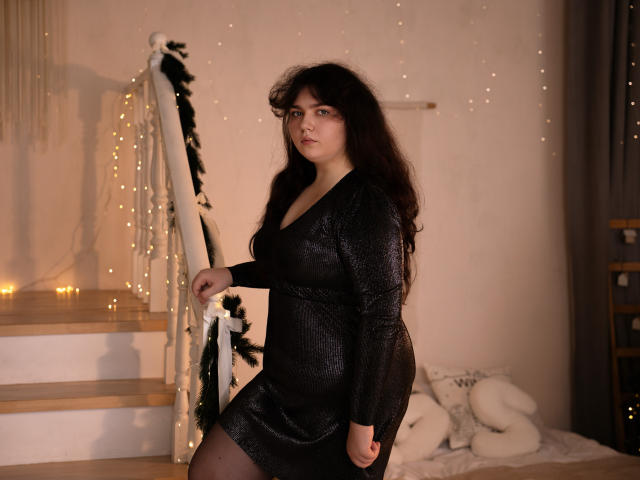 NeonilaLisova with dark curly hair, wearing a shiny black dress, standing on wooden stairs, holding the railing, looking at the camera, soft light, candles in the background.