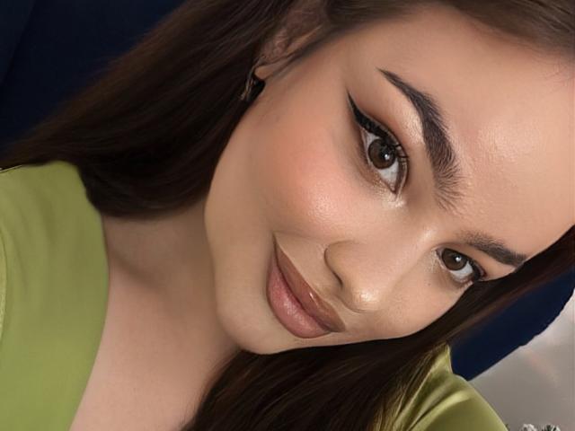 LilyCartere has shiny brown eyes, thick black eyebrows, and full, glossy brown lips. She's wearing a green top, with her brown hair partially visible. Her skin is smooth.