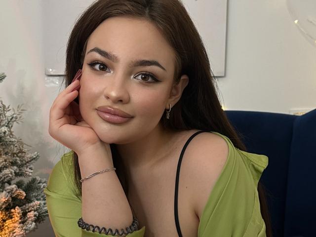 LilyCartere with her pouty lips, brown eyes, and glossy pink lipstick, wearing a green top with a black strap, hand on her cheek, looking cute.