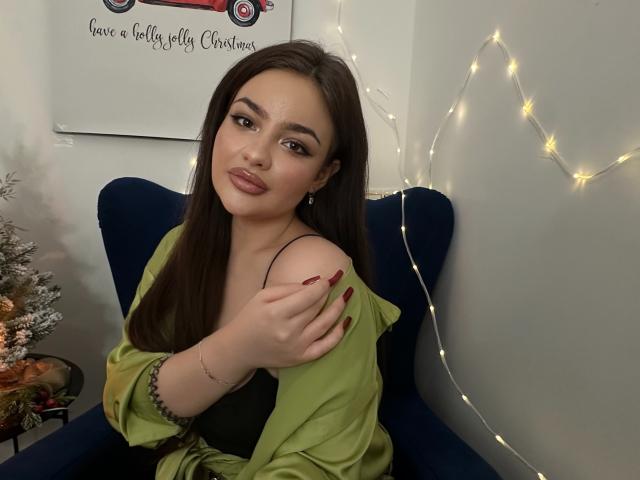 LilyCartere with long brown hair, green off-shoulder shirt, black bra, red nail polish, and a bracelet, sitting in a blue chair, looking at the camera.