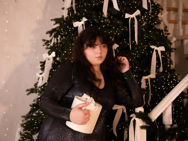 NeonilaLisova with dark hair, wearing a sparkly black dress, holding a white gift box with a bow, standing next to a decorated Christmas tree with white ribbons.