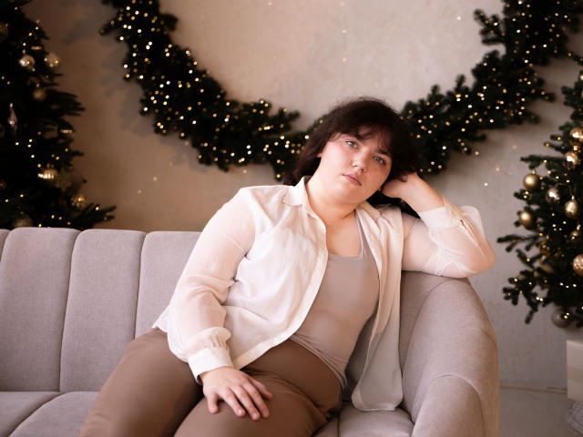 NeonilaLisova, a fair-skinned woman with dark, wavy hair, sits on a light grey sofa. She wears a loose, white, button-down shirt over a beige tank top and brown pants. Her right arm rests on the sofa's back while her left hand touches her head. She has a relaxed, slightly bored expression. Christmas decorations with green garlands and small yellow lights are in the background. The image is a photograph with a soft, warm light. Watermark in the bottom right corner.