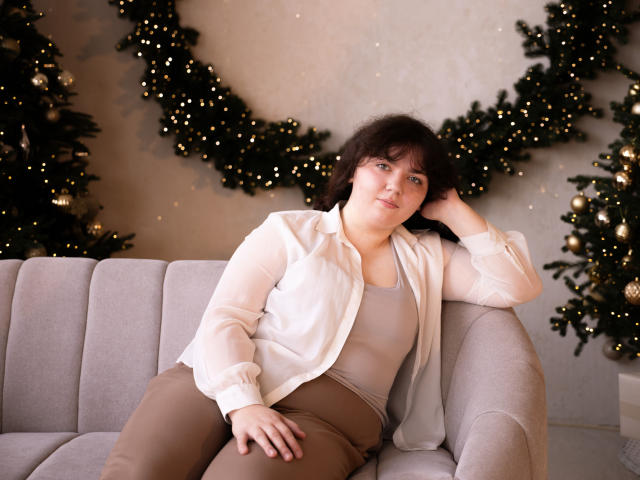 NeonilaLisova with short brown hair, blue eyes, and light skin, wearing a white shirt and beige pants, sits on a gray sofa, hand on head, looking relaxed.
