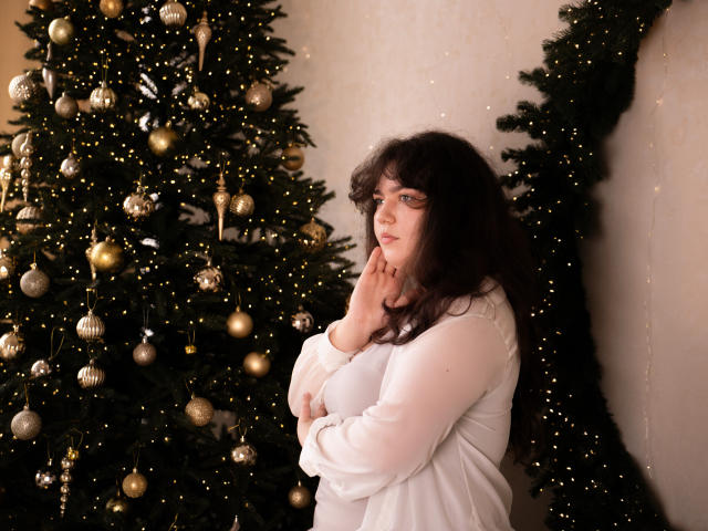NeonilaLisova with curly dark hair, white long-sleeve shirt, standing near a decorated Christmas tree, hands near face, looking slightly sad, boobs partially visible, soft lighting.