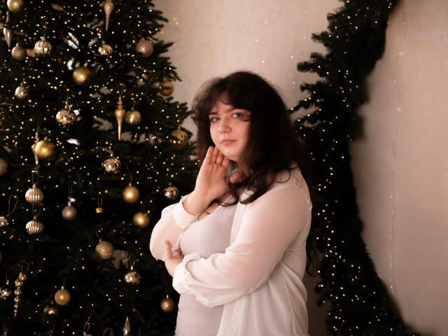 NeonilaLisova with wavy dark hair, wearing a white long-sleeve top, hands near her face, standing in front of a decorated Christmas tree with gold ornaments and lights.