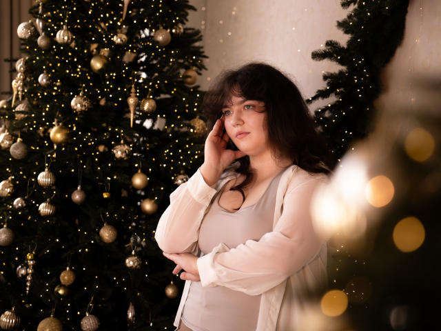 NeonilaLisova with curly dark hair, wearing a white cardigan and light gray top, touching her cheek, standing in front of a decorated Christmas tree with golden lights and ornaments.