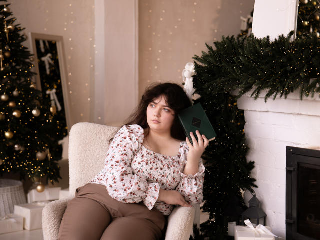 NeonilaLisova sits in a beige armchair with her left hand holding a green book. She has dark brown, wavy hair and wears a floral blouse with puffed sleeves and brown pants. She looks to the right with a neutral expression. The background includes a decorated Christmas tree with yellow lights and ornaments, and a white brick fireplace. The image has a soft, warm light and a cozy, festive atmosphere. The composition is balanced with NeonilaLisova slightly off-center.