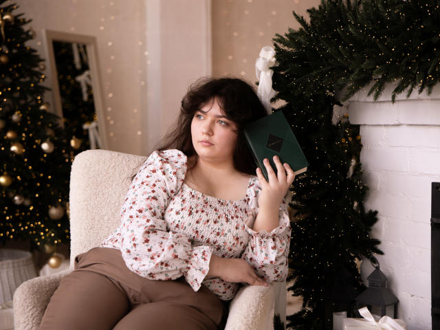 NeonilaLisova sits in a plush, cream-colored armchair. She has medium-length, wavy brown hair and a slightly worried expression. She wears a floral blouse with long sleeves and brown pants. She holds a dark green book in her right hand, resting it on her shoulder. Behind her, a decorated Christmas tree with gold ornaments and fairy lights is partially visible. A white brick fireplace is to her right, adorned with greenery. The composition uses warm, soft lighting, highlighting her and the festive decor. Her posture is relaxed yet slightly anxious.