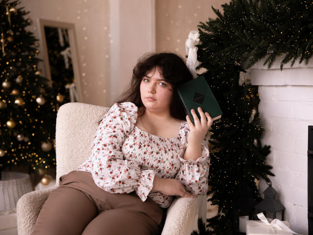 NeonilaLisova, a plus-sized woman with long dark hair, sits in a cream-colored armchair. She wears a floral blouse and brown pants. She holds a green book with a black "A" in her right hand. Her left arm rests on the armchair. She's positioned near a decorated Christmas tree with green foliage and yellow ornaments. The white brick fireplace in the background has a small lantern on the right. The image uses soft lighting, highlighting her facial expression and the textures of her clothing and chair.