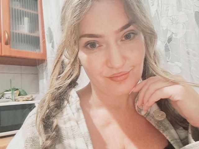CarmenAutumn with wavy brown hair, green eyes, and light skin, wearing a grey, button-up, knit top, has a subtle smile and rests her hand near her chin.