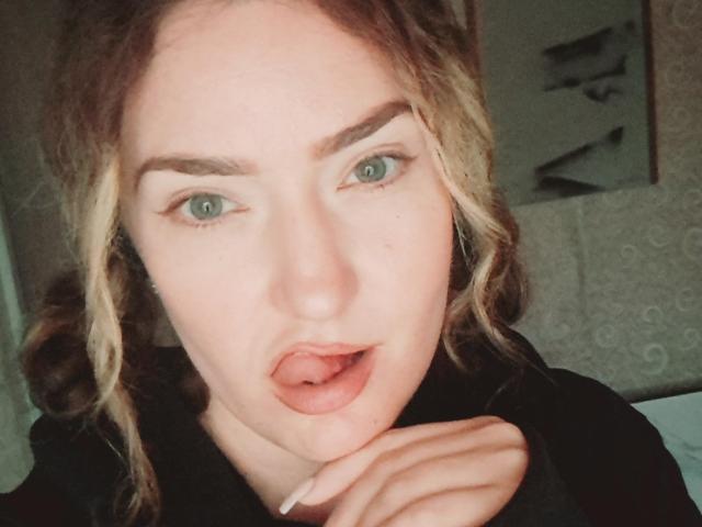 CarmenAutumn has blue eyes, slightly open mouth with tongue touching upper lip, light brown wavy hair, and is touching her chin with her right hand. She's wearing a black hoodie.