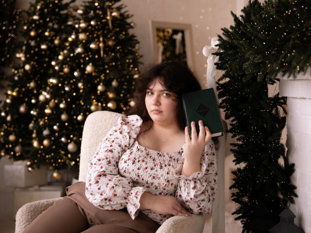 NeonilaLisova sits in a cream-colored armchair, holding a green book with a black emblem. She has fair skin, dark wavy hair, and wears a floral-patterned, off-shoulder blouse with brown pants. Her right arm rests on the armchair's armrest. Behind her, a decorated Christmas tree with golden lights and ornaments fills the background. A green garland is draped over a white fireplace to her right. The image has a warm, cozy feel, with soft lighting and a focus on NeonilaLisova's contemplative expression.