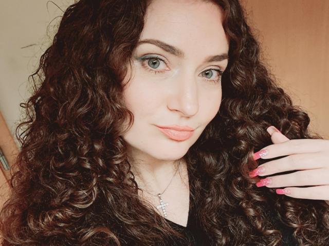 CarmenAutumn has curly brown hair, green eyes, pink lips, and pink nails with a silver necklace. She's looking at the camera with a slight smile.