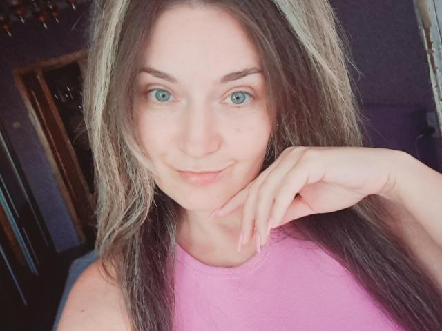 CarmenAutumn with green eyes, smiling, wearing a pink top, her hand near her chin, long brown hair with blonde highlights, slight pink nose, soft light on her face.