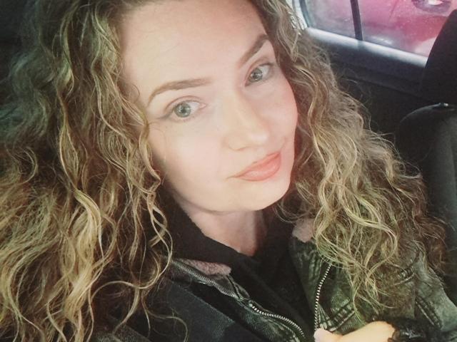 CarmenAutumn has curly blonde hair, green eyes, and full pink lips. She's wearing a green jacket and black shirt, sitting in a car with arms crossed.