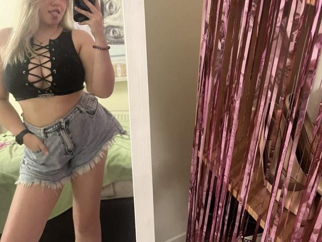 UniqueLady with blonde hair, black lace-up crop top, showing her tits, and frayed denim shorts, sticking out her tongue in a mirror selfie. Pink curtain on the right.