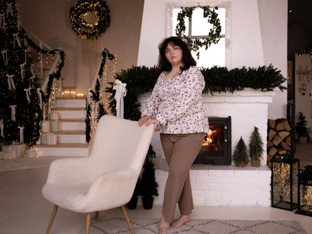 NeonilaLisova stands barefoot in a cozy room, leaning on a white, textured armchair. She has dark, wavy hair and wears a floral-patterned blouse with brown pants. A white brick fireplace with a lit fire is behind her, adorned with greenery and small Christmas trees. The room is warmly lit with string lights on the stairs and a wreath on the wall. Her expression is neutral. The image is a photograph.