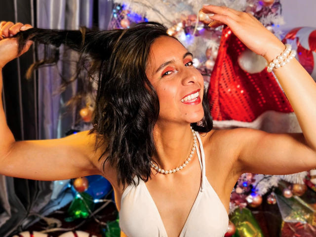 MiaAmoree with black wavy hair, holding it, smiling, wearing a white halter top, pearl necklace, smiling with teeth showing, Christmas tree and red hat in the background.