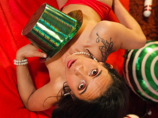 MiaAmoree with shiny green cup over tits, wearing red lingerie and pearl necklace, lying on red blanket, looking up with a sexy smile, black tattoo on right shoulder.