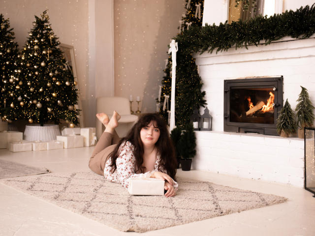 NeonilaLisova lies on a beige rug in a cozy room, reading a book. She has long, dark brown hair and wears a floral shirt with brown pants. Her feet are up, and she rests on her elbows. Two Christmas trees with gold and white ornaments are in the background, and a white fireplace with a green garland and lit fire is to the right. The scene is warm and festive.