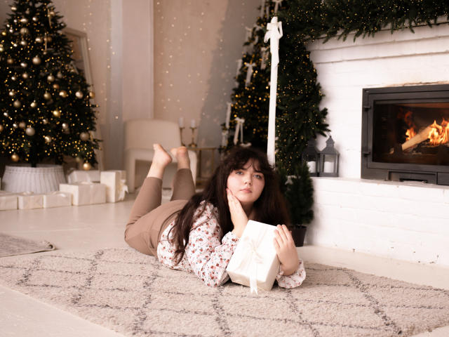 NeonilaLisova lies on a beige carpet in a cozy room, facing the camera with a relaxed expression. She has long dark hair, wears a floral long-sleeve top, and beige pants. Her head rests on her right hand, and she holds a white gift box with both hands. Two decorated Christmas trees and a lit fireplace are in the background. The room has soft, warm lighting with string lights on the wall. Her feet are crossed in the air. The image has a warm, inviting feel.