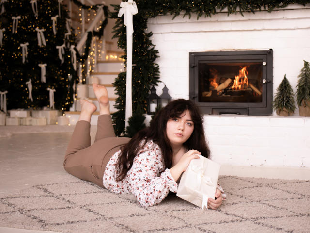 NeonilaLisova, a young woman with long dark hair, lies on a beige carpet in front of a white brick fireplace with a lit fire. She wears a floral blouse and brown pants, holding a white gift box. Her feet are up, and she looks slightly to the side. The background includes a decorated Christmas tree and lit string lights. The image has a cozy, festive feel. Her expression is neutral, and the lighting is soft.