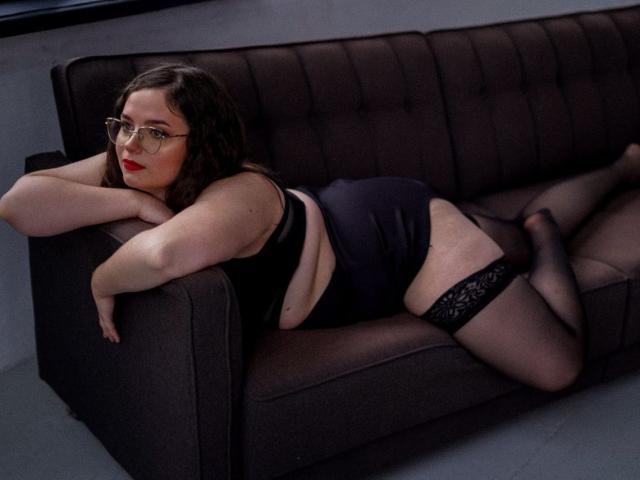 SophieDreams is lying on a brown sofa, wearing black lingerie, thigh-high stockings, and glasses, with her ass up and her tits down, looking at the camera.