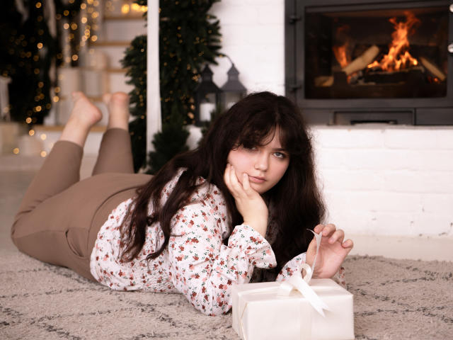 NeonilaLisova lies on a carpet, wearing a floral shirt and brown pants, with a gift box in front, her ass up, looking at the camera, fire burning in the background.