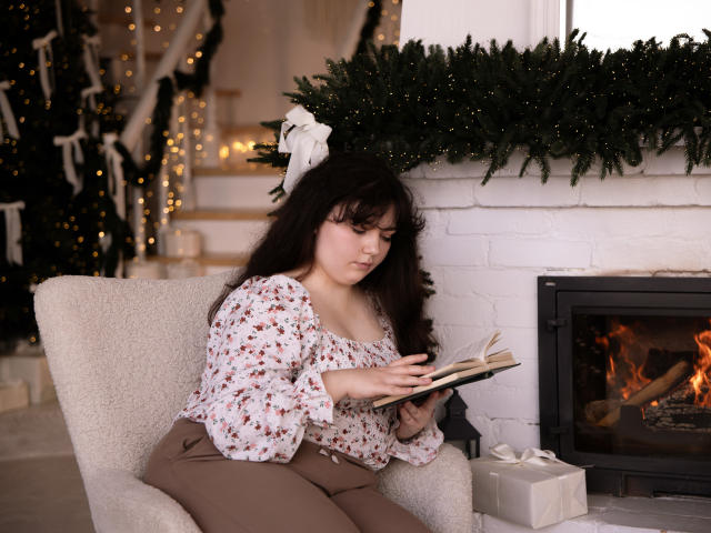 NeonilaLisova with curly brown hair, wearing a floral blouse and brown pants, reading a book, sitting in a white armchair, by a fireplace with a green garland.