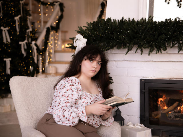 NeonilaLisova, a young woman with long dark hair, sits in a beige armchair reading a book. She wears a floral blouse with puffed sleeves and brown pants. Her expression is focused. A white bow is in her hair. She's in a cozy room with a white brick fireplace, green garland, and a lit Christmas tree in the background. The fireplace has a small fire. She holds a book open in her hands. The image is warmly lit.