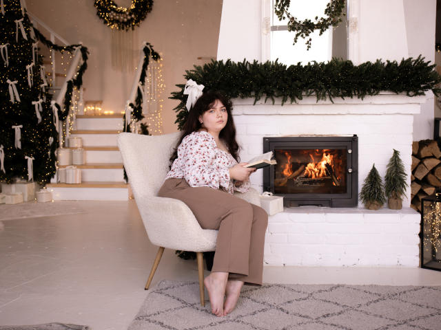 NeonilaLisova, with curly dark hair and a white floral blouse, sits barefoot in a cream armchair by a lit fireplace. She wears brown pants and reads a book, her expression calm. The fireplace is centered with green garland above, and small Christmas trees flank it. The room has white stairs with string lights and a wreath on the wall. The floor is light wood with a gray patterned rug. The image is cozy and festive.