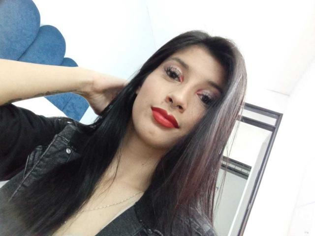 AlisKooper with long black hair, red lipstick, and a black denim jacket, looking at the camera with a slight smile, hand behind head, in a white room.