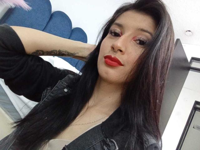AlisKooper has long black hair, red lipstick, and a tattoo on her right arm. She's wearing a black jacket, looking at the camera with a slight pout.