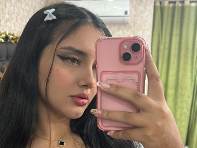 AmaliaDalton with long black hair, pink lipstick, and bold eyeliner, holding a pink phone, showing off her nails, with a small blue hair clip, looking at the camera.