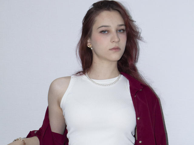 TorriAustin has red hair, blue eyes, and is wearing a white tank top with a red velvet jacket, pearl necklace, and gold earrings, looking slightly annoyed.
