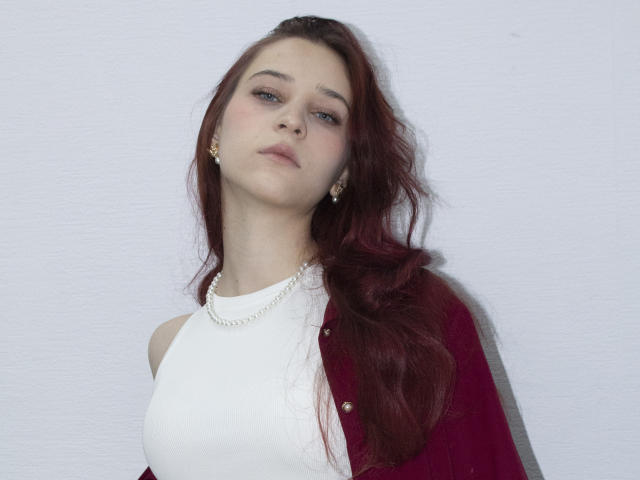 TorriAustin with red hair, white sleeveless top, pearl necklace, and red cardigan, looking slightly down, wearing small gold earrings, pale skin, subtle makeup, and a neutral expression.