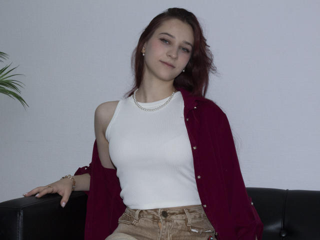 TorriAustin with reddish-brown hair, white tank top, red open shirt, tan pants, and necklace, sitting on black couch, looking slightly to the side, small earrings, subtle smile.