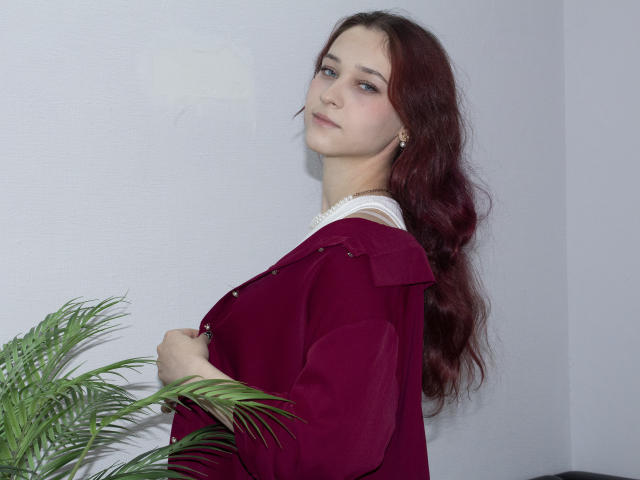 TorriAustin with long red hair, wearing a deep red jacket over a white top, holding it slightly open, standing next to a green plant, looking sideways with a slight smile.
