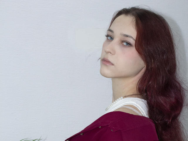 TorriAustin with red hair, pale skin, and blue eyes, wearing a white top and red shirt, looking over shoulder, subtle makeup, necklace, soft expression, upper body shot.